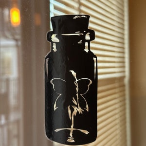 May include: A black silhouette of a jar with a butterfly inside, on a glass surface with a blurred background.