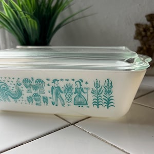 May include: A rectangular, vintage Pyrex baking dish with a clear glass lid. The dish is off-white with a teal-colored, stylized farm scene design around the sides. The design includes a rooster, baskets, and figures of people.