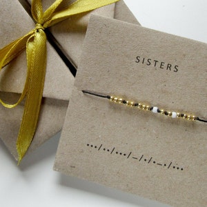 Può includere: Una scatola regalo di carta marrone con un nastro giallo legato in un fiocco. La scatola è su una superficie bianca. Un piccolo biglietto con la parola "SISTERS" stampata su di esso è accanto alla scatola. Un sottile filo nero con perline dorate e bianche è sul biglietto.