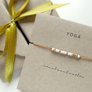 Può includere: Un braccialetto fatto a mano con perline bianche e dorate infilate su un cordoncino arancione, presentato su un biglietto di carta kraft. Il biglietto recita "YOGA" e presenta il codice Morse. Un nastro dorato è legato attorno a un pacco di carta kraft.