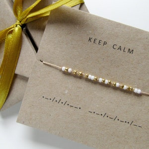 Può includere: Un biglietto marrone con la scritta "KEEP CALM" stampata su di esso. Un braccialetto di perline bianche e dorate è esposto sul biglietto.
