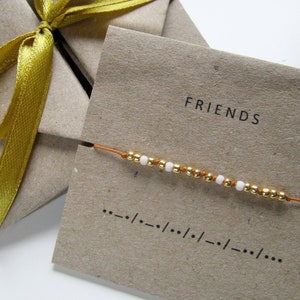 Può includere: Un cartoncino marrone con la parola "FRIENDS" stampata su di esso. Un sottile filo marrone con piccole perline dorate e bianche è infilato attraverso i fori nel cartoncino.