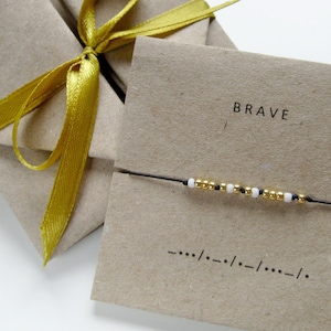 Op de afbeelding: Een eenvoudige armband met gouden en witte kralen op een zwart koord. De armband is verpakt in een bruine papieren doos met een gouden lint en een kaart met de tekst "BRAVE".