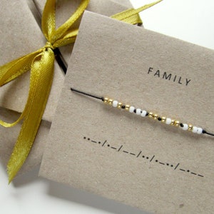 Può includere: Un biglietto marrone con la parola "FAMILY" stampata su di esso. Uno spago nero con perline bianche e dorate è infilato attraverso il biglietto. Il biglietto ha anche una serie di punti e trattini, che è il codice Morse per la parola "FAMILY".