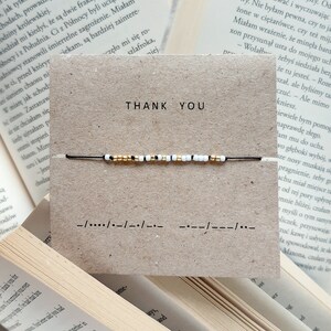 Può includere: Un braccialetto di corda nero con perline bianche e dorate. Il braccialetto è su una carta marrone con le parole "Grazie" e un messaggio in codice Morse.