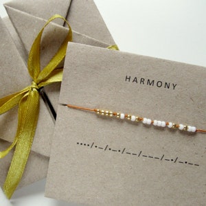 Può includere: Un cartoncino di carta marrone con la parola "HARMONY" stampata in nero. Un sottile filo marrone con perline dorate e bianche è mostrato sulla carta. La carta presenta anche una linea di punti e trattini, che rappresentano il codice Morse.