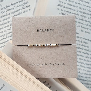 Può includere: Un braccialetto di corda nero con perline bianche e dorate. Il braccialetto è su una carta marrone con la parola "BALANCE" stampata in lettere nere. La carta ha anche una linea di trattini e le parole "for the" stampate su di essa.