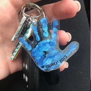 May include: A clear acrylic keychain in the shape of a handprint, painted with blue paint. The keychain is attached to a keyring with keys and a car remote. The name "Oliver 2017" is written on the keychain.