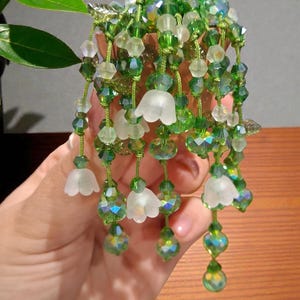 May include: A handcrafted floral hair accessory with strands of green and clear crystal beads, and white flower charms. The design incorporates various shades of green and clear beads, creating a shimmering, elegant effect. A decorative hair piece.
