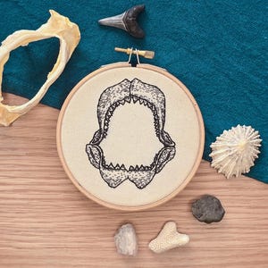 May include: An embroidery hoop with a black and white shark jaw design, alongside a shark tooth, bone, and various small rocks and shells. The background is a teal cloth and a wooden surface.