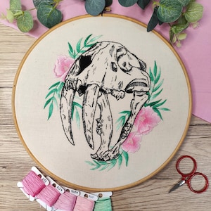 May include: Embroidered hoop art featuring a detailed black and white saber-toothed cat skull surrounded by green leaves and pink flowers. The hoop is on a wooden surface with spools of pink and green embroidery floss.