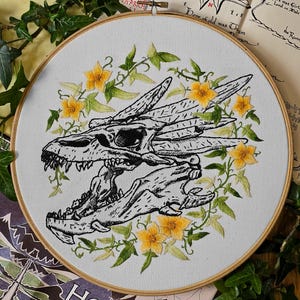 May include: An embroidered dragon skull design in a wooden hoop. The black and white skull is surrounded by yellow flowers and green leaves. The embroidery is on a light gray fabric. The word "HOBBIT" is visible.