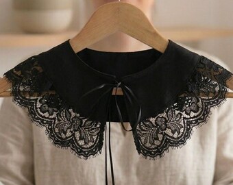 Puede incluir: Un cuello postizo negro con una base de tela lisa y un borde ancho de encaje floral intrincado. El cuello se asegura en la parte delantera con una cinta negra fina que cuelga, creando un acento decorativo en este accesorio de vestir.