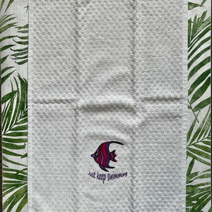 May include: White kitchen towel with a red, purple and white fish design and the text "Just keep swimming".