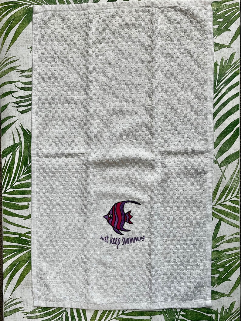 May include: White kitchen towel with a red, purple and white fish design and the text "Just keep swimming".