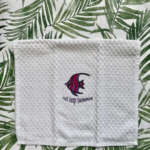 May include: White kitchen towel with an embroidered purple and red fish and the text "Just keep swimming".