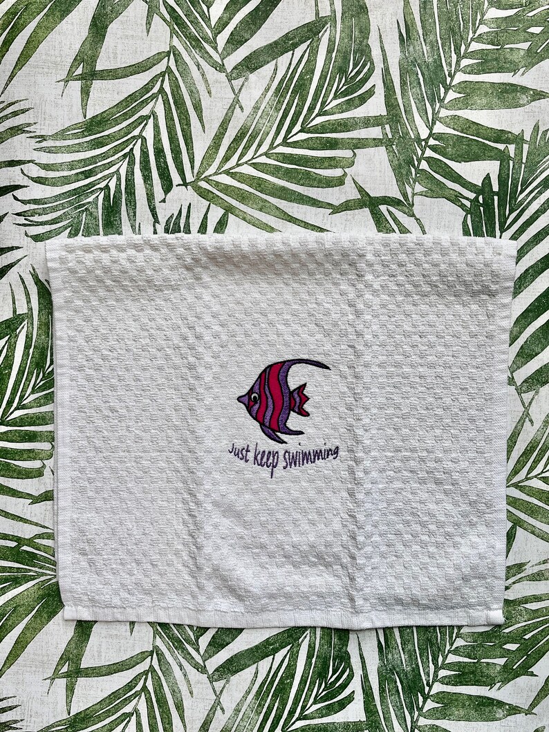 May include: White kitchen towel with an embroidered purple and red fish and the text "Just keep swimming".