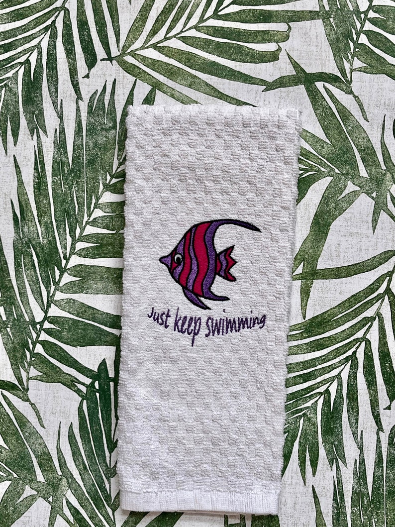 May include: White kitchen towel with an embroidered purple and red fish and the text "Just keep swimming".