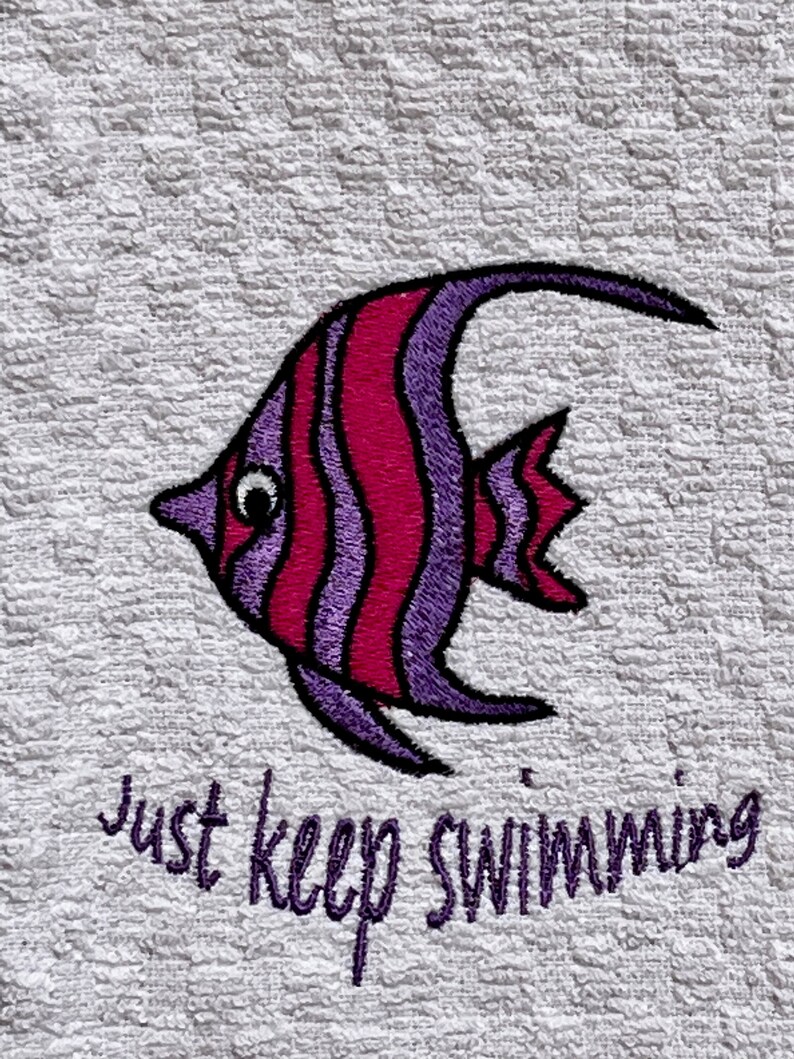 May include: A white towel with a purple and pink embroidered fish and the text "just keep swimming".