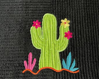 Puede incluir: Una toalla de tela negra texturizada con un cactus verde bordado de líneas verticales. El cactus tiene dos flores rosas y una amarilla. En la base, hay pequeños acentos de plantas rosas y azules sobre una línea curva de color naranja.