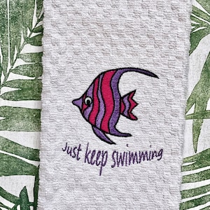 May include: White kitchen towel with an embroidered purple and pink fish and the text "Just keep swimming".