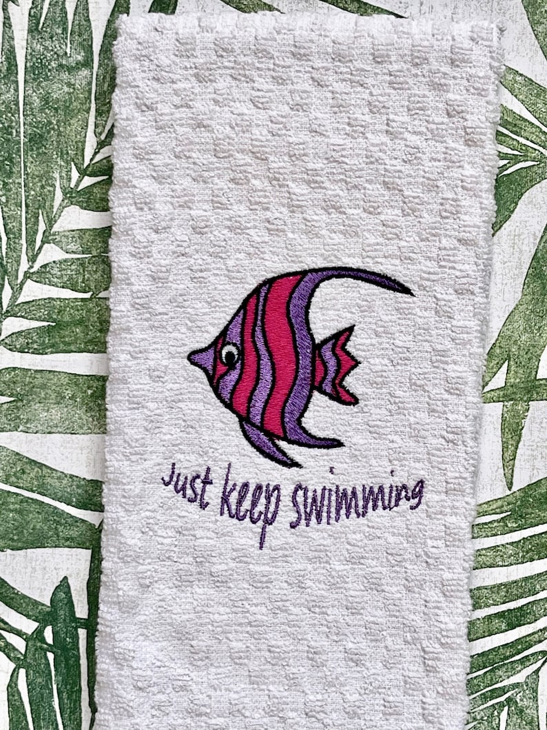 May include: White kitchen towel with an embroidered purple and pink fish and the text "Just keep swimming".
