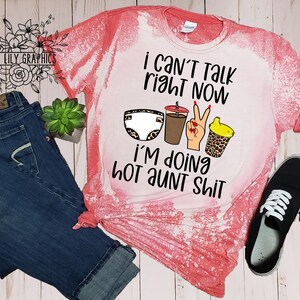 May include: A pink bleached t-shirt with the text "I can't talk right now I'm doing hot aunt shit" and a graphic of a diaper, a cup of coffee, a hand with peace sign, and a sippy cup.