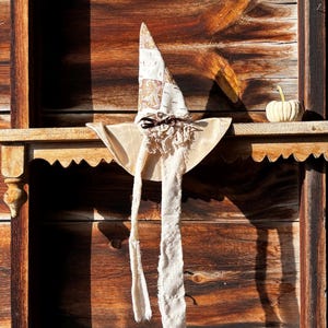 Può includere: Un cappello a punta con un cono patchwork nei toni del beige e del bianco con motivi paisley, una tesa larga beige e un fiocco di nastro marrone scuro. Due lunghe strisce di tessuto bianco sporco sfilacciate pendono dalla base.