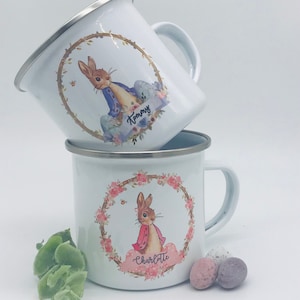 May include: Two white enamel mugs with silver rims. Each mug features a floral wreath design with a cartoon rabbit. One mug has the name "Tommy" and the other has "Charlotte". The mugs are stacked on top of each other.