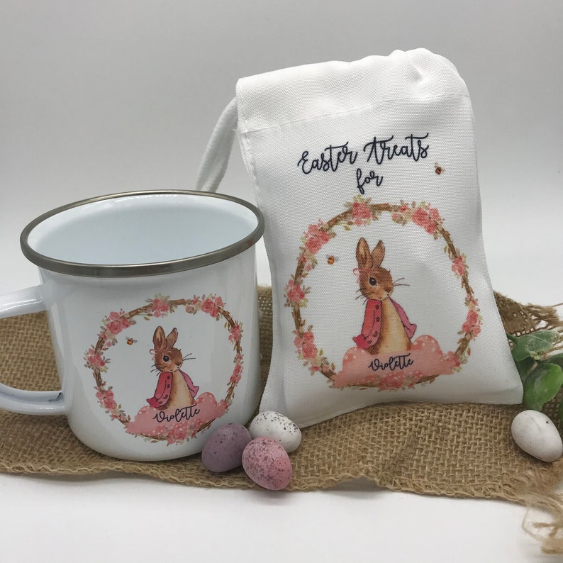 May include: White enamel mug and drawstring bag with a bunny design. The mug and bag feature a bunny wearing a pink coat, surrounded by a floral wreath. The bag reads "Easter Treats for" with the name "Violette" below the bunny.