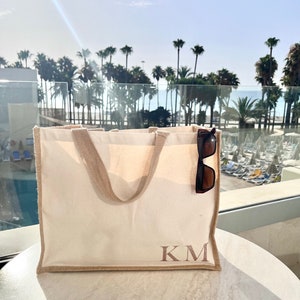 May include: A white canvas tote bag with brown trim and the initials "KM" embroidered in brown on the front. Sunglasses are tucked into the top of the bag.