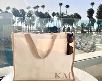 Op de afbeelding: Een witte canvas tote bag met bruine bies en de initialen "KM" in bruin geborduurd op de voorkant. Een zonnebril is in de bovenkant van de tas gestoken.