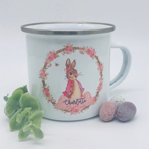 May include: White enamel mug with a silver rim and handle. The mug features a floral wreath design with a cartoon rabbit wearing a pink jacket. The name "Charlotte" is written on a pink cloud. Easter-themed.