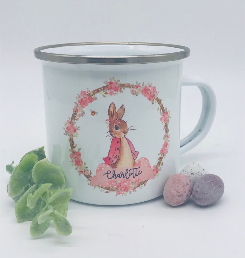May include: White enamel mug with a silver rim and handle. The mug features a floral wreath design with a cartoon rabbit wearing a pink jacket. The name "Charlotte" is written on a pink cloud. Easter-themed.