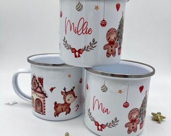 May include: Three white enamel mugs with Christmas designs. The mugs have red and green designs with a gingerbread man, a Christmas tree, a reindeer, and a house. The mugs are personalized with the names "Millie" and "Mia".