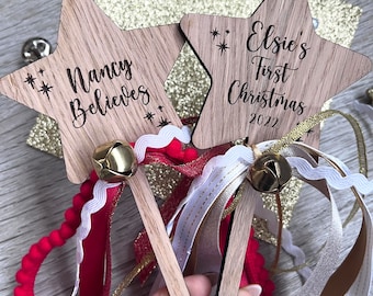 Könnte beinhalten: Zwei sternförmige Holzornamente mit Glöckchen und Bändern. Ein Ornament trägt die Aufschrift "Nancy Believes" und das andere "Elsie's First Christmas 2022".
