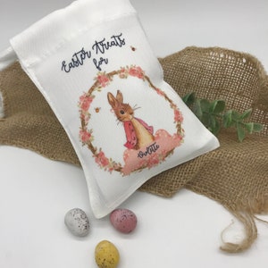 May include: White drawstring bag with the text "Easter Treats for" and a watercolor illustration of a bunny in a pink coat inside a floral wreath. The name "Violette" is written below the bunny. Three speckled Easter eggs are in the foreground.