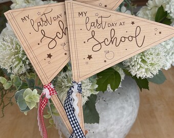 May include: Two wooden pennant flags with the text "My last day at School" in a cursive font. The flags have star and apple illustrations. Red and white, and blue and white checkered ribbons are attached to the flag handles.