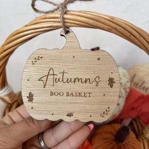 May include: A wooden pumpkin-shaped tag with the text "Autumn's Boo Basket" engraved on it. The tag is hanging from a wicker basket.