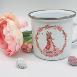 May include: White enamel mug with a silver rim, featuring a watercolor illustration of a bunny in a pink jacket, surrounded by a floral wreath. The name "Charlotte" is written below the bunny. A pink flower and speckled eggs are next to the mug.