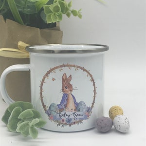 May include: White enamel mug with a silver rim and handle. The mug features a watercolor illustration of a rabbit in a blue jacket, surrounded by a floral wreath and Easter eggs. The name "Finley Beau" is written on the mug.