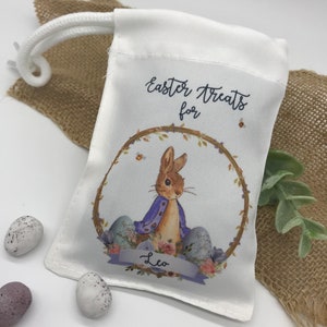 May include: White drawstring bag with an Easter theme. The bag features a watercolor illustration of a bunny in a purple jacket inside a wreath, with the text "Easter Treats for" and the name "Leo". Three speckled Easter eggs are next to the bag.