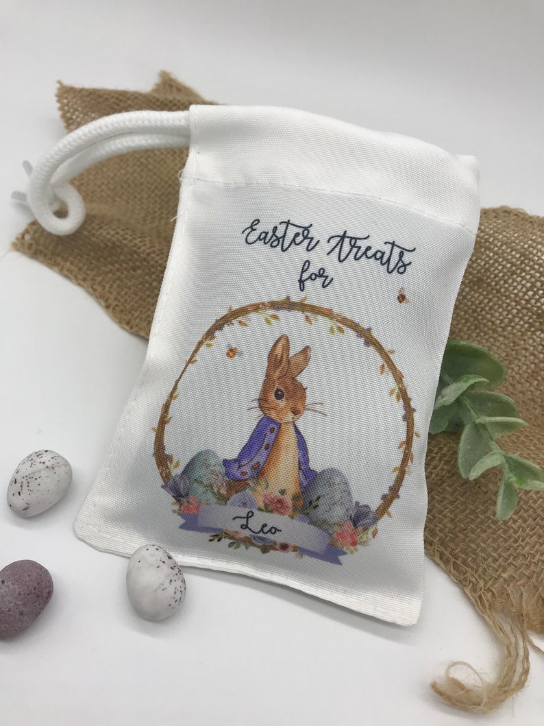 May include: White drawstring bag with an Easter theme. The bag features a watercolor illustration of a bunny in a purple jacket inside a wreath, with the text "Easter Treats for" and the name "Leo". Three speckled Easter eggs are next to the bag.