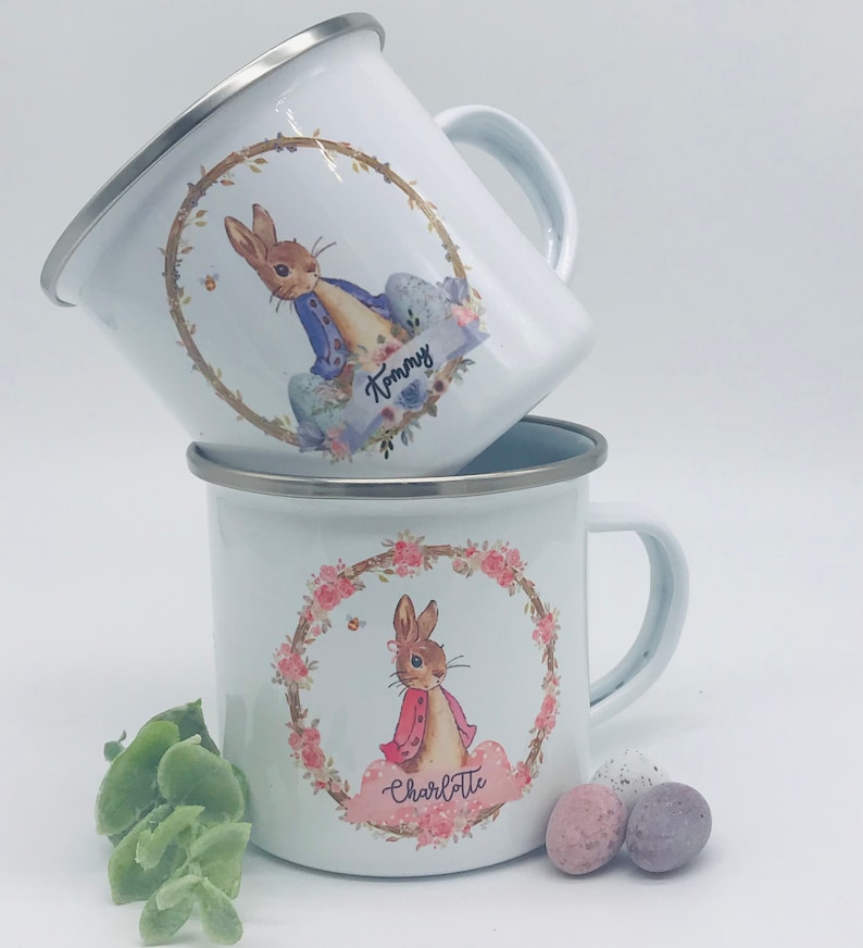 May include: Two white enamel mugs with silver rims. Each mug features a floral wreath design with a cartoon rabbit. One mug has the name "Tommy" and the other has "Charlotte". The mugs are stacked on top of each other.