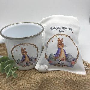 May include: White enamel mug and drawstring bag featuring a bunny illustration in a floral wreath. The bag reads "Easter Treats for" and includes the name "Leo". A small, speckled egg sits nearby, suggesting an Easter theme.