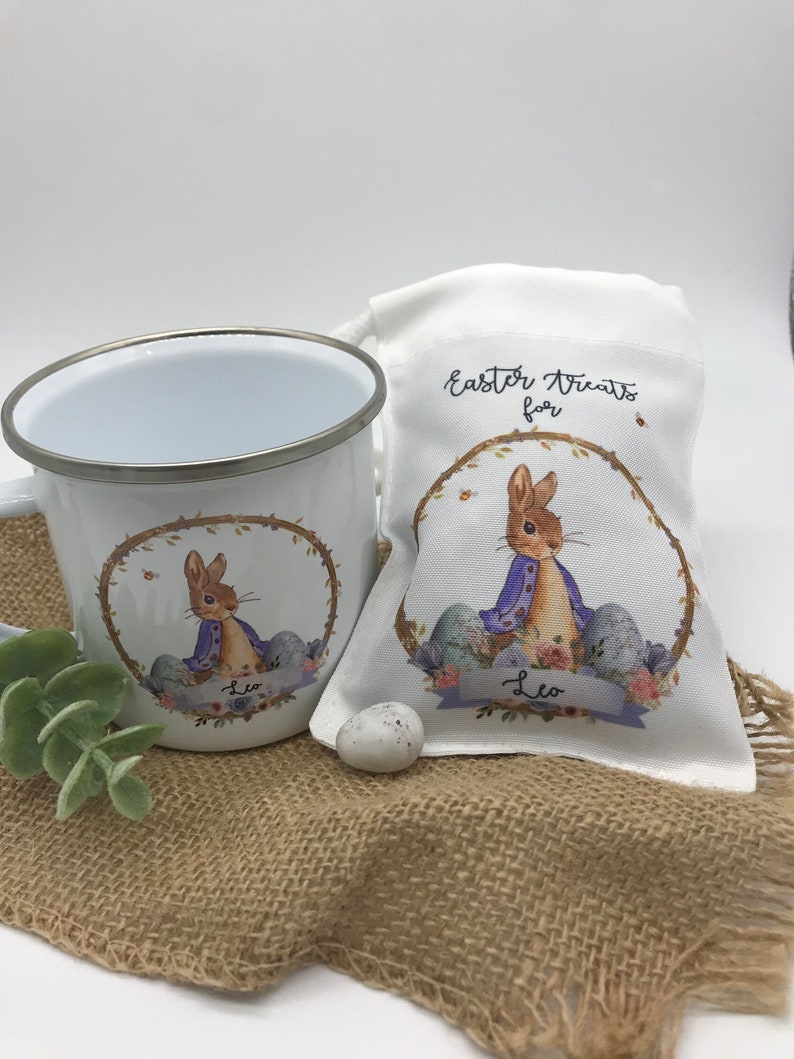 May include: White enamel mug and drawstring bag featuring a bunny illustration in a floral wreath. The bag reads "Easter Treats for" and includes the name "Leo". A small, speckled egg sits nearby, suggesting an Easter theme.