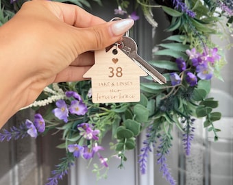 Può includere: Portachiavi in legno a forma di casa con il testo "Jake & Lindsey our forever home" e il numero 38. Il portachiavi è tenuto da una mano su uno sfondo di fiori viola.
