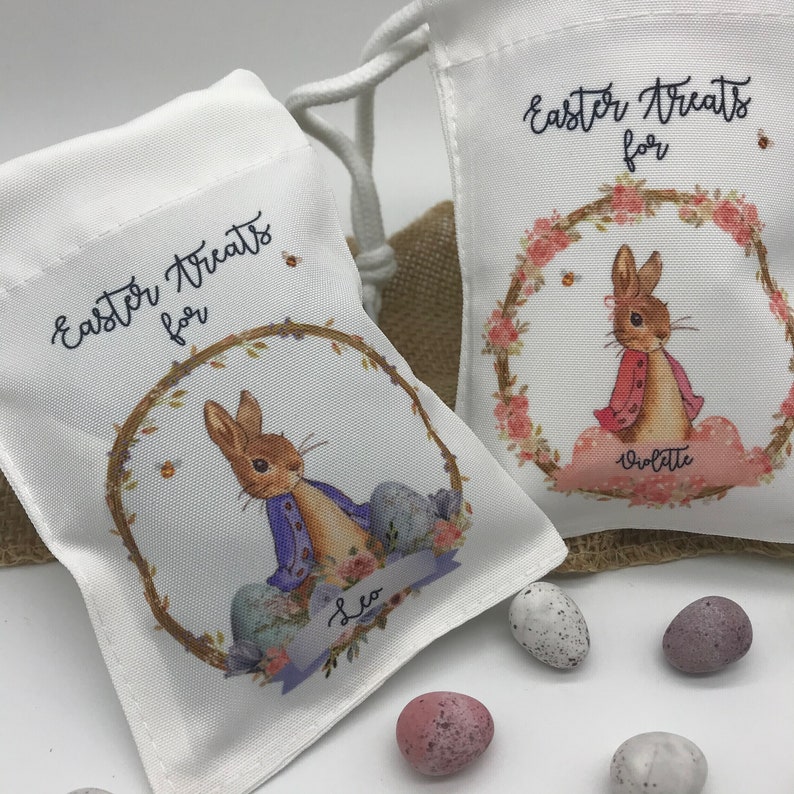 May include: Two white drawstring bags with the text "Easter Treats for" and a bunny illustration. One bag has the name "Leo" and the other "Violette". The bags are decorated with floral wreaths and Easter eggs. Several speckled eggs are scattered around the bags.
