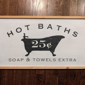 May include: A white wooden sign with black lettering that reads "HOT BATHS 25¢ SOAP & TOWELS EXTRA". The sign features a black silhouette of a bathtub.