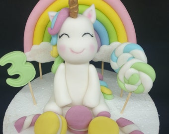 May include: A white unicorn cake topper with a rainbow, a cloud, and a lollipop. The cake is decorated with pink and yellow macarons and the name "KAYA" is written in pink icing.